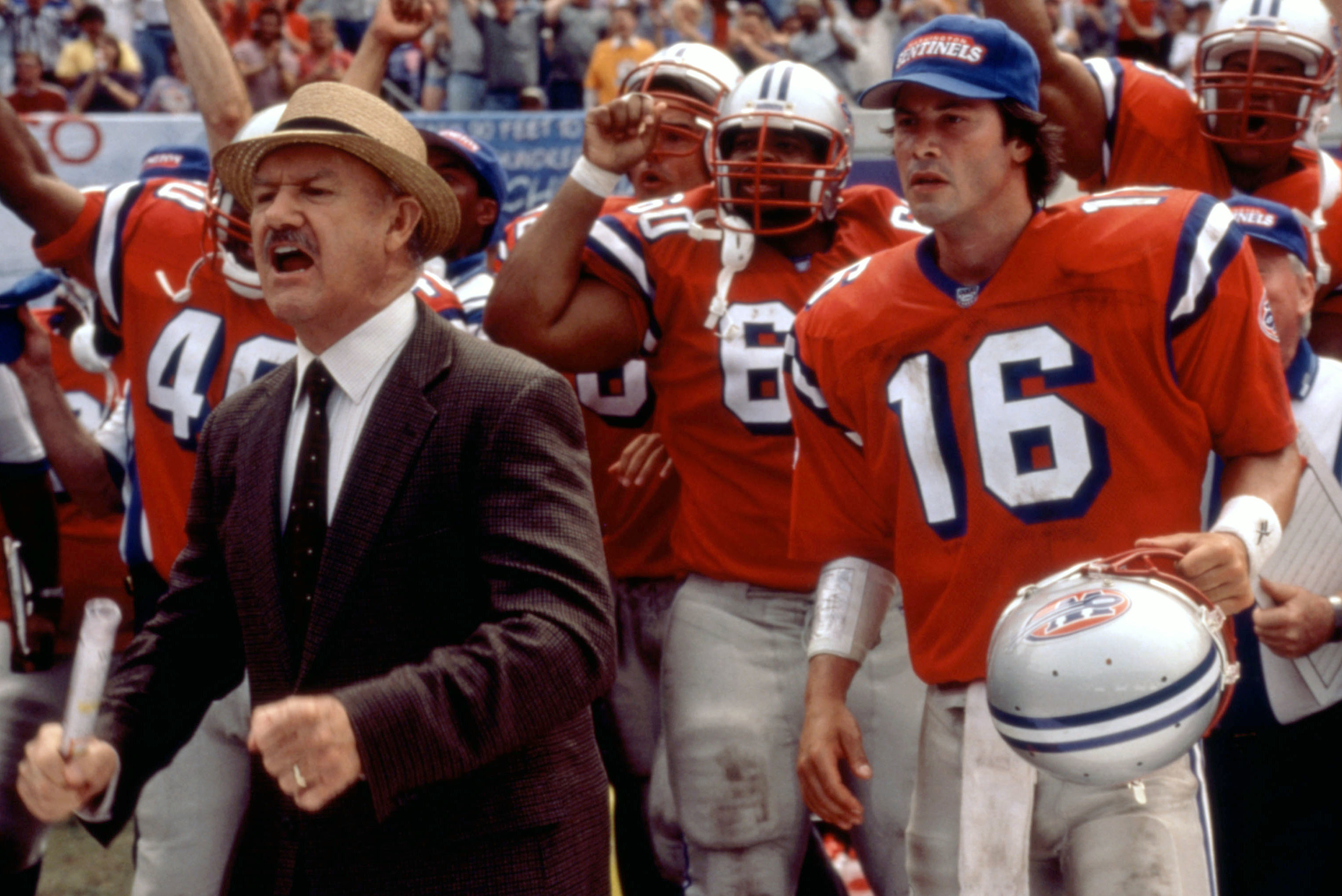 Gene Hackman in a suit and Keanu Reeves in football gear on the sidelines with a cheering football team wearing helmets and jerseys in the background