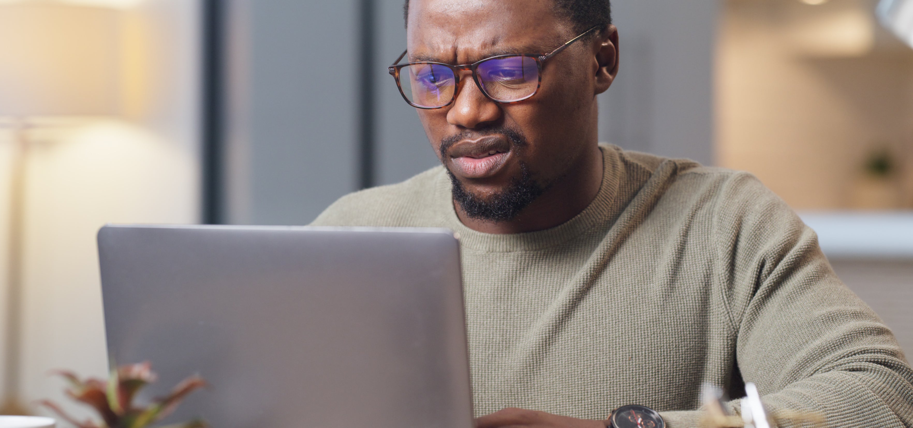 A person with glasses and a beard, wearing a sweater, looks intently at a laptop screen in a modern workspace