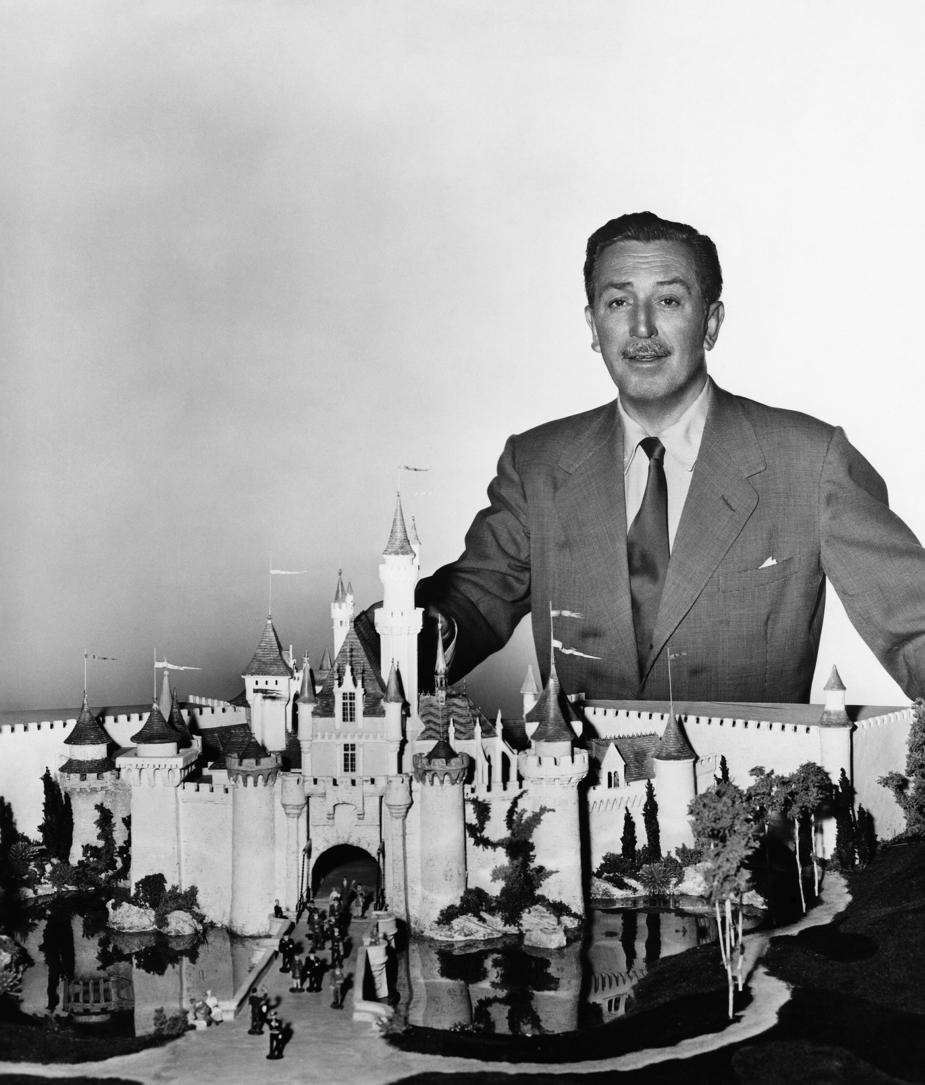 Walt Disney stands behind a model of Disneyland's Sleeping Beauty Castle, wearing a suit and tie, with a proud expression on his face
