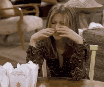 Jennifer Aniston dramatically holds her nose while appearing to sniff her hair at a dining table with Chinese takeout boxes in front of her