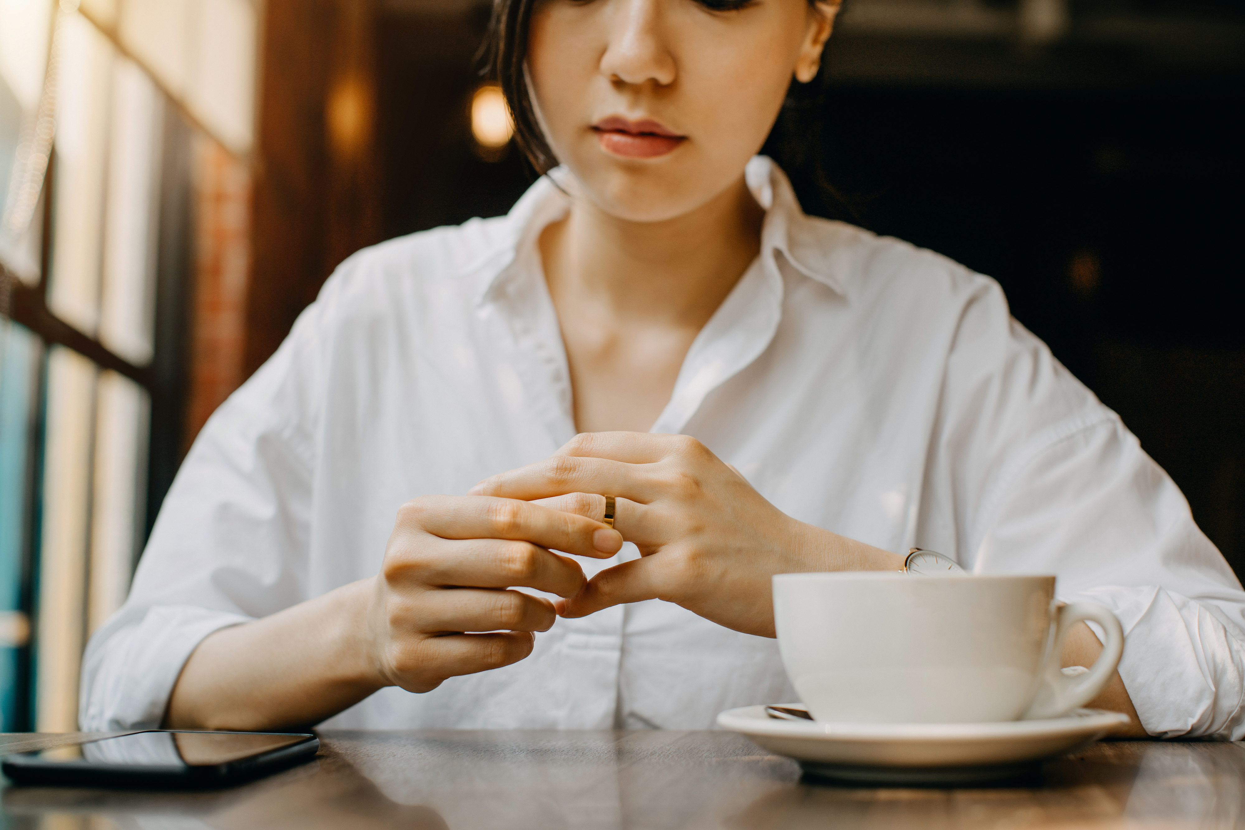 A woman sits at a table wearing a white shirt, holding a ring with a cup of coffee in front of her. Her face is partially visible and she appears thoughtful