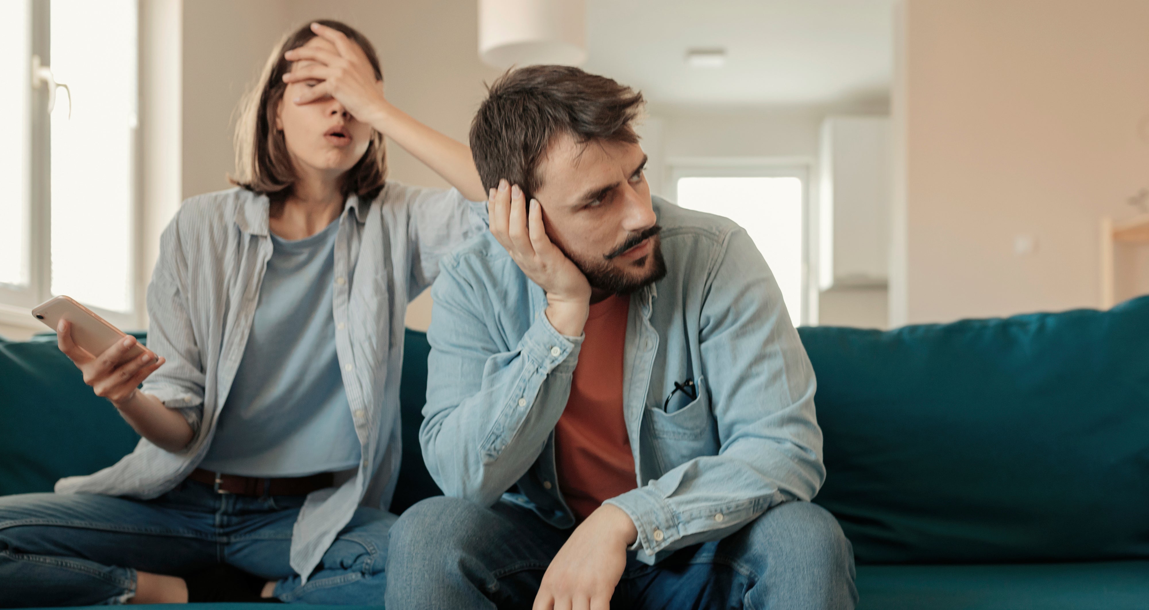 A woman sitting on a sofa playfully covers a man's eyes while he looks contemplative, resting his head on his hand. Both are casually dressed