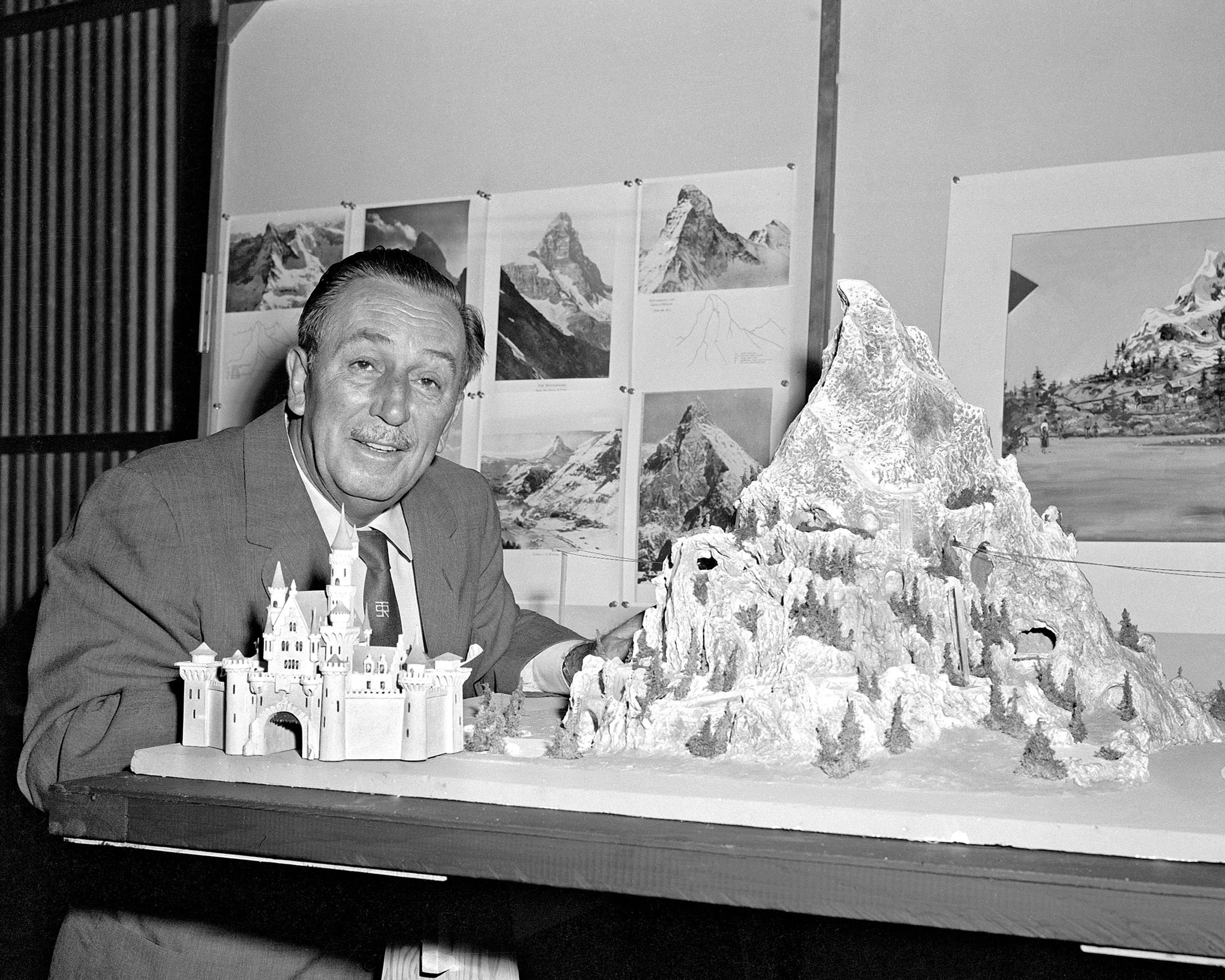 Walt Disney is leaning on a table displaying a model of a castle and a mountainous landscape, likely a theme park attraction