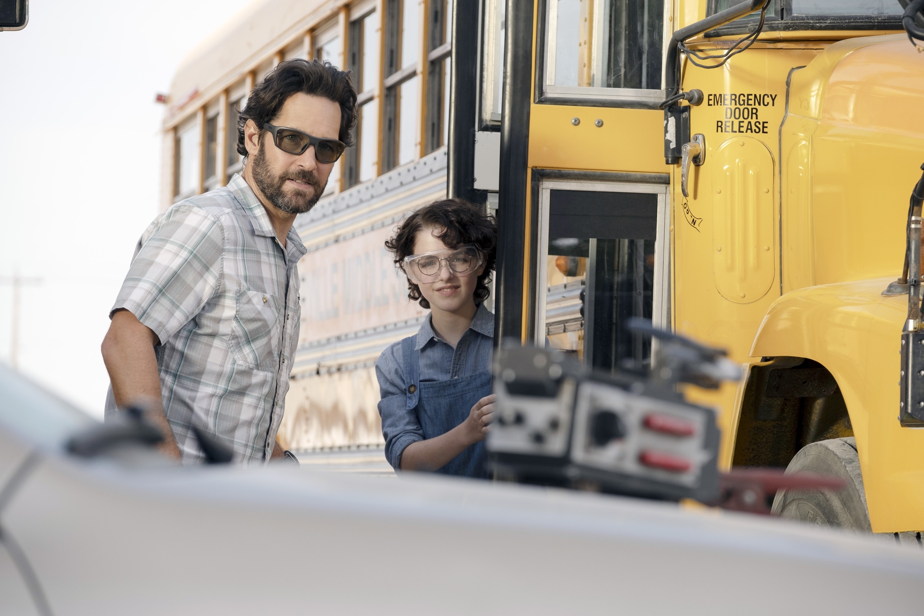 Paul Rudd and Finn Wolfhard stand beside a school bus, looking towards a parked car