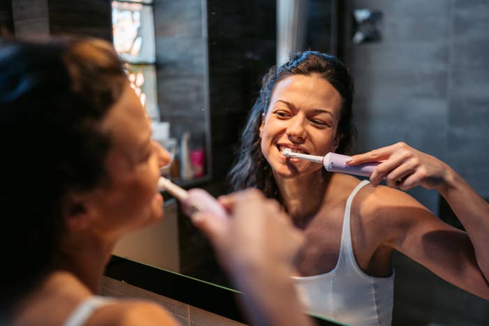 A woman brushes her teeth with an electric toothbrush while looking in the bathroom mirror