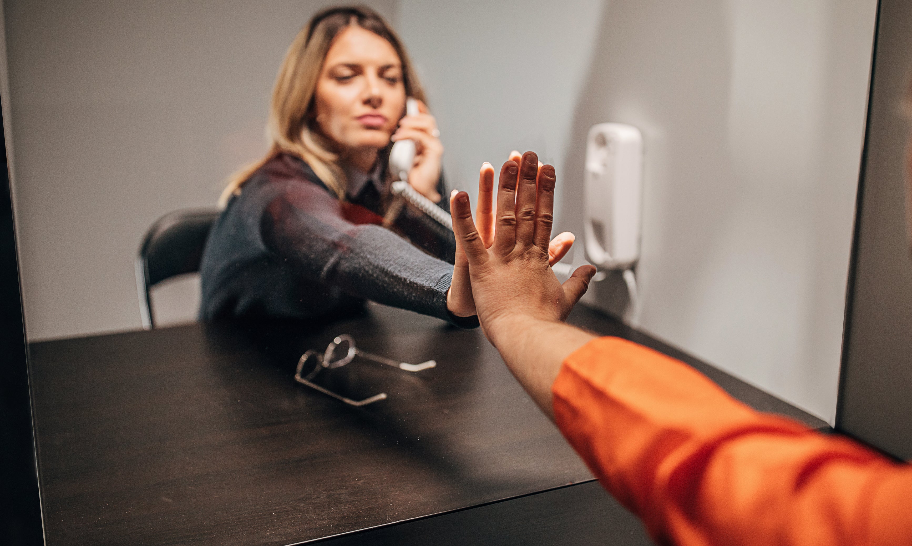A woman and an incarcerated man touch hands through glass while speaking using a phone in a visitation room