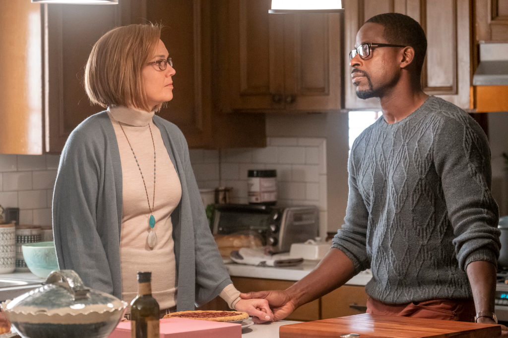 Mandy Moore and Sterling K. Brown in a kitchen scene. Mandy wears a turtleneck and cardigan, while Sterling wears a cable-knit sweater. They appear serious and engaged in conversation