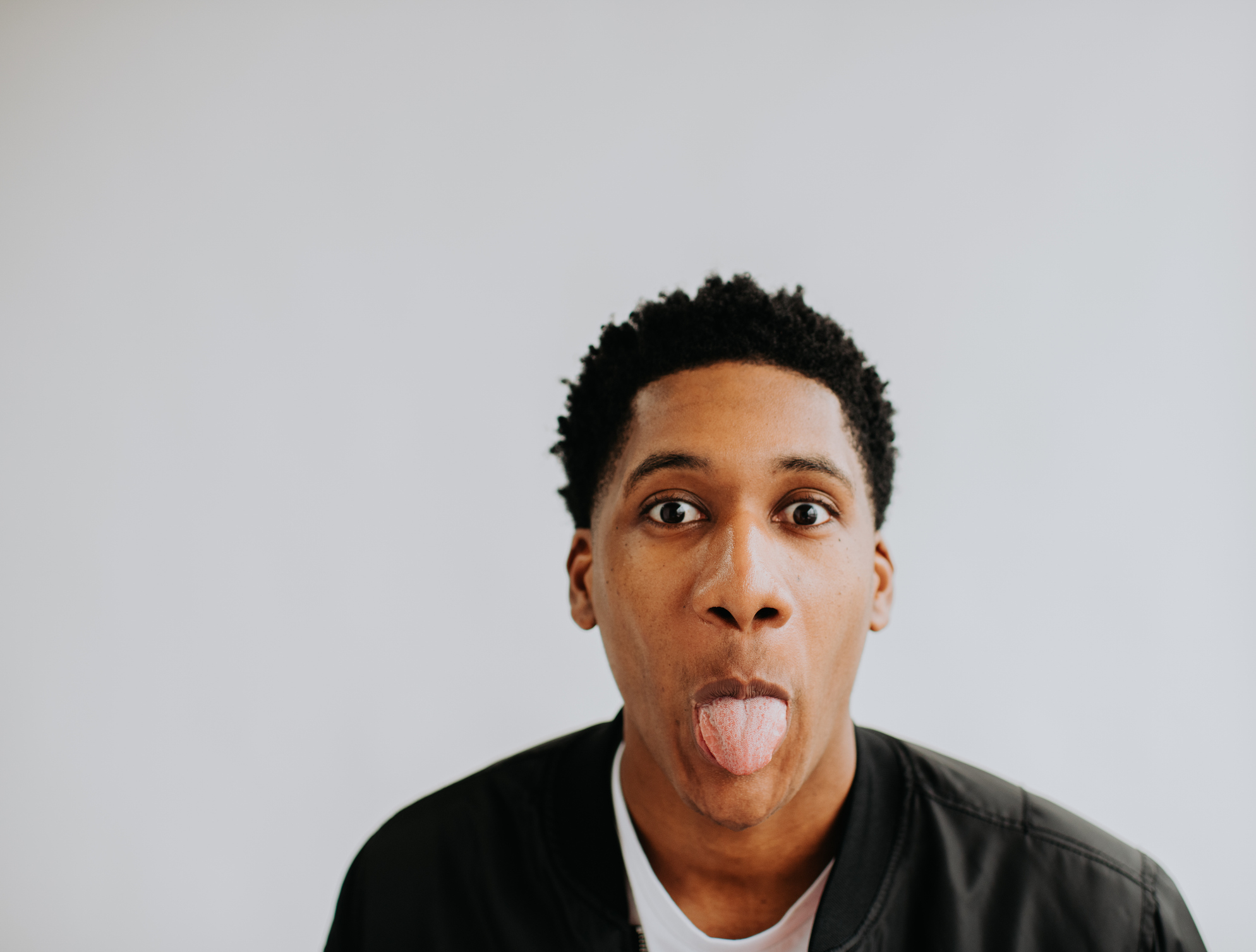 A person with short curly hair makes a playful face, sticking out their tongue, against a plain background