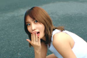 Twice band member covering her mouth with one hand while crouching on a street. They are wearing a sleeveless top and have shoulder-length hair and look surprised.