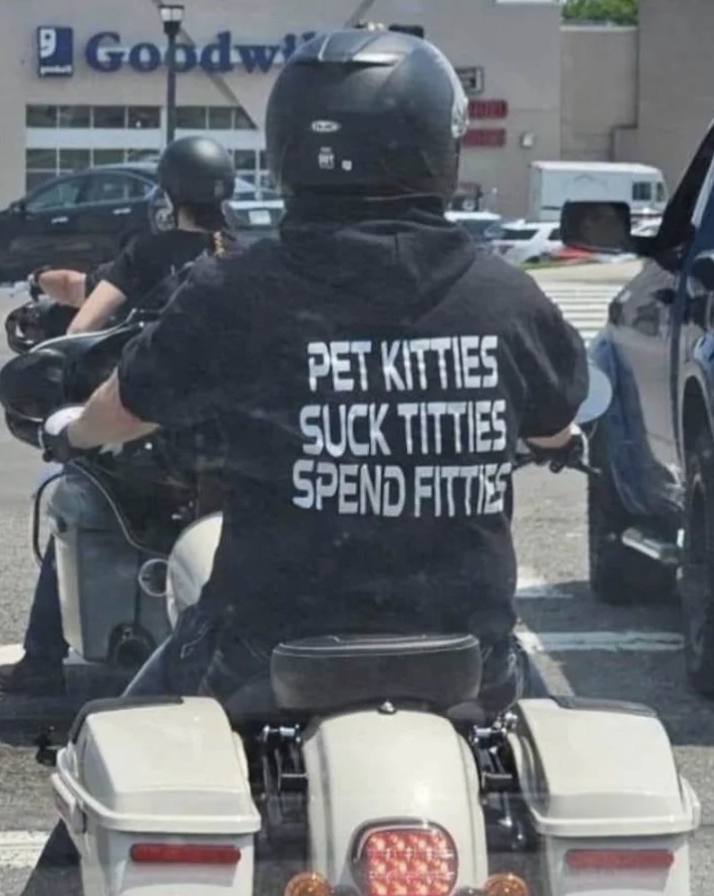 A person wearing a black helmet and a T-shirt that reads "PET KITTIES SUCK TITTIES SPEND FITTIES" is riding a motorcycle in a parking lot