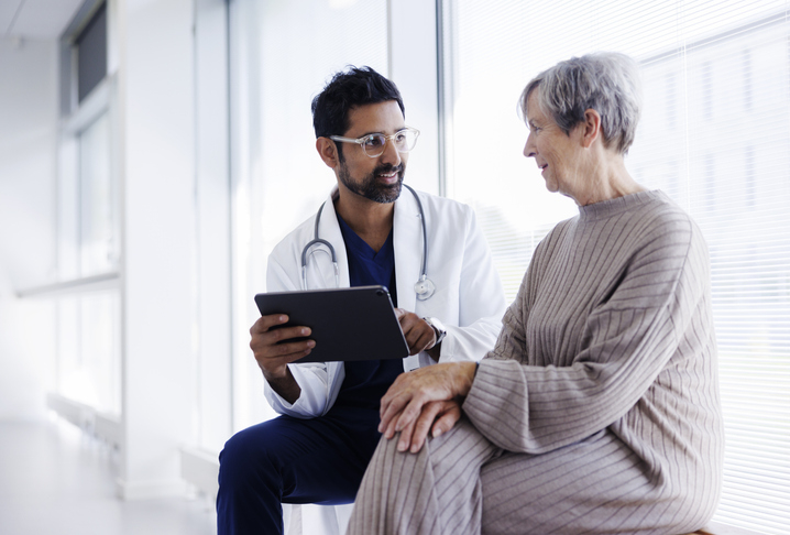 A doctor holding a tablet talks to an older woman seated beside him in a well-lit room