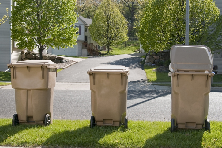 Three tan garbage bins lined up on a residential street, with houses and green trees in the background. One bin's lid is partially open