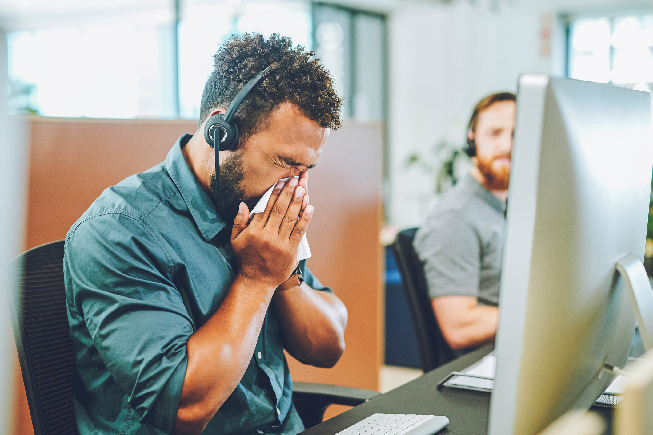 A man at a desk in an office sneezes into a tissue while wearing headphones, with a co-worker in the background