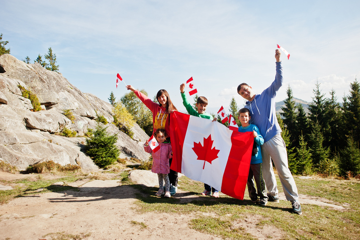 A family of five, two adults and three children, waves Canadian flags and holds a large Canadian flag while standing outdoors in a mountainous area
