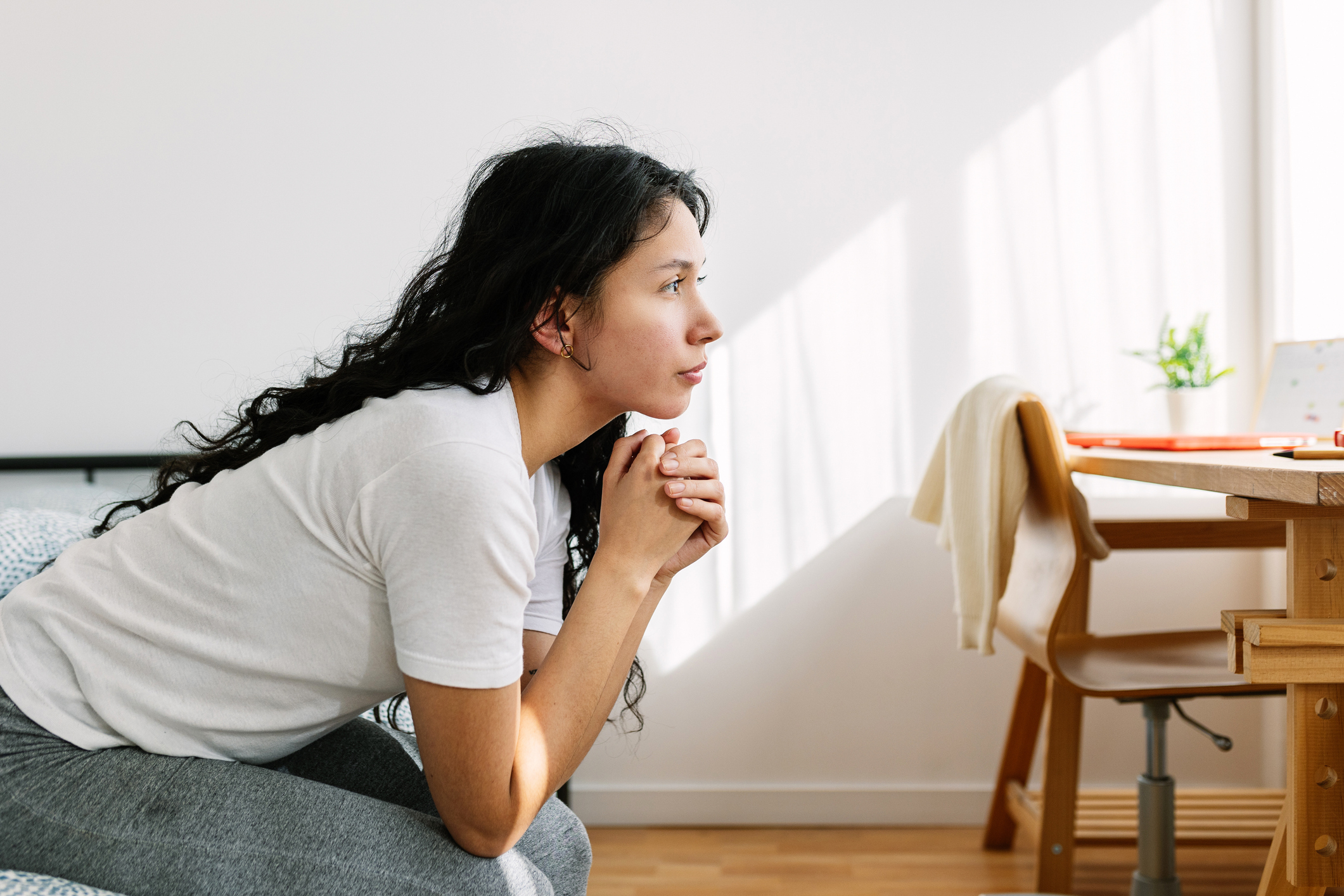 A woman with long dark hair sits on a bed, leaning forward with her hands clasped, looking towards a sunlit window. A desk with items is in the background