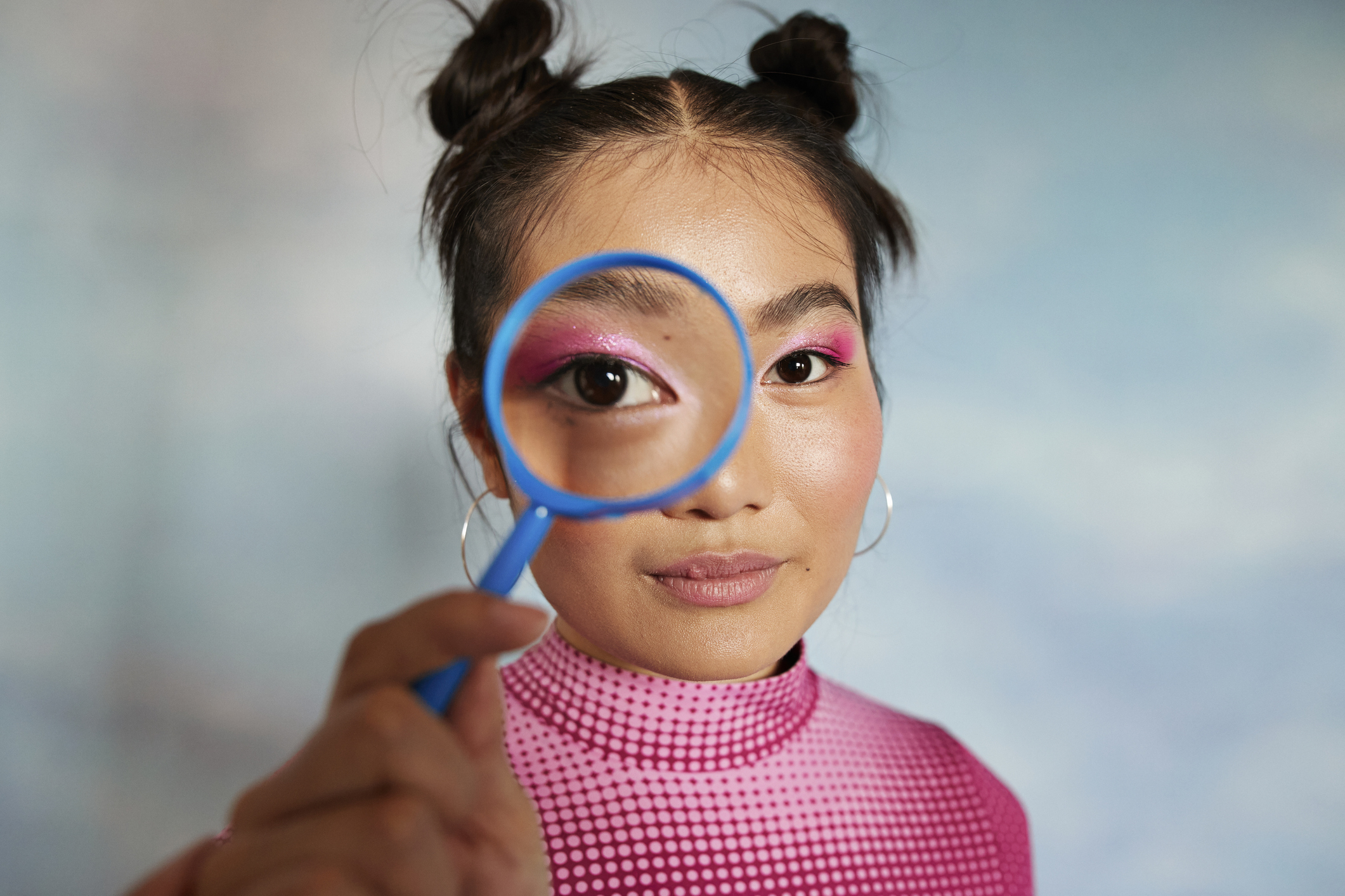 A young woman with two top buns peers through a magnifying glass held to her eye. She wears hoop earrings and a vibrant, patterned top