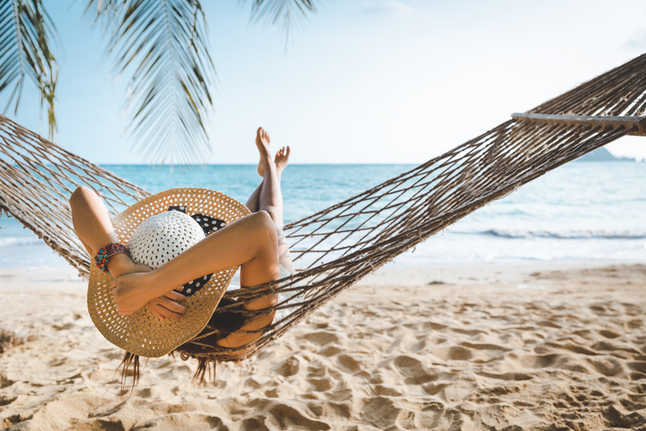 Person relaxing in a hammock on a beach, wearing a sun hat. Beach and ocean in the background
