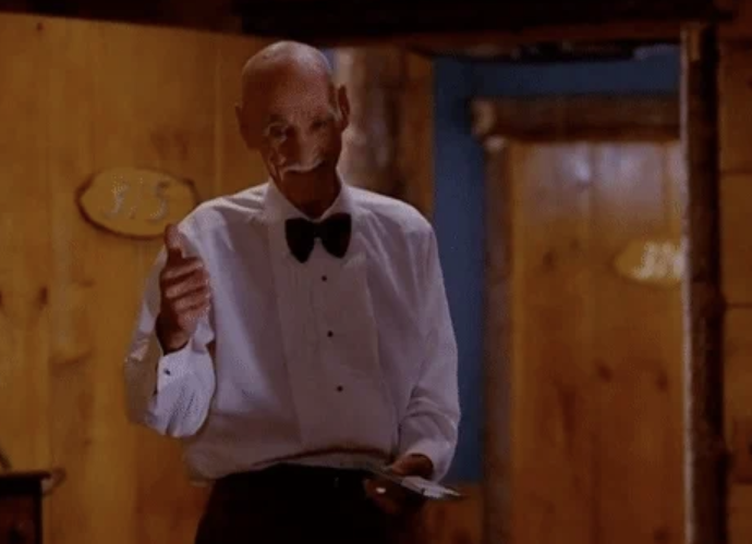 An older man in a white shirt and black bow tie gives a thumbs-up while holding a piece of paper in what appears to be a rustic setting
