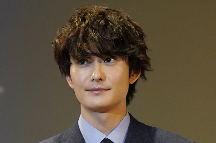 綾田将生がダブルブレストのスーツを着て、胸に赤いリボンをつけて立っている。