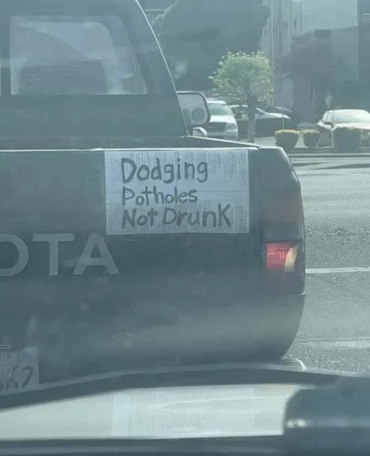 A sign on the back of a Toyota truck reads, "Dodging Potholes Not Drunk." The truck is waiting at a traffic light