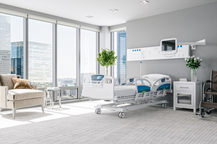 Modern hospital room with a large window, a hospital bed, a beige armchair, a medical monitor, and a small cabinet with medical equipment