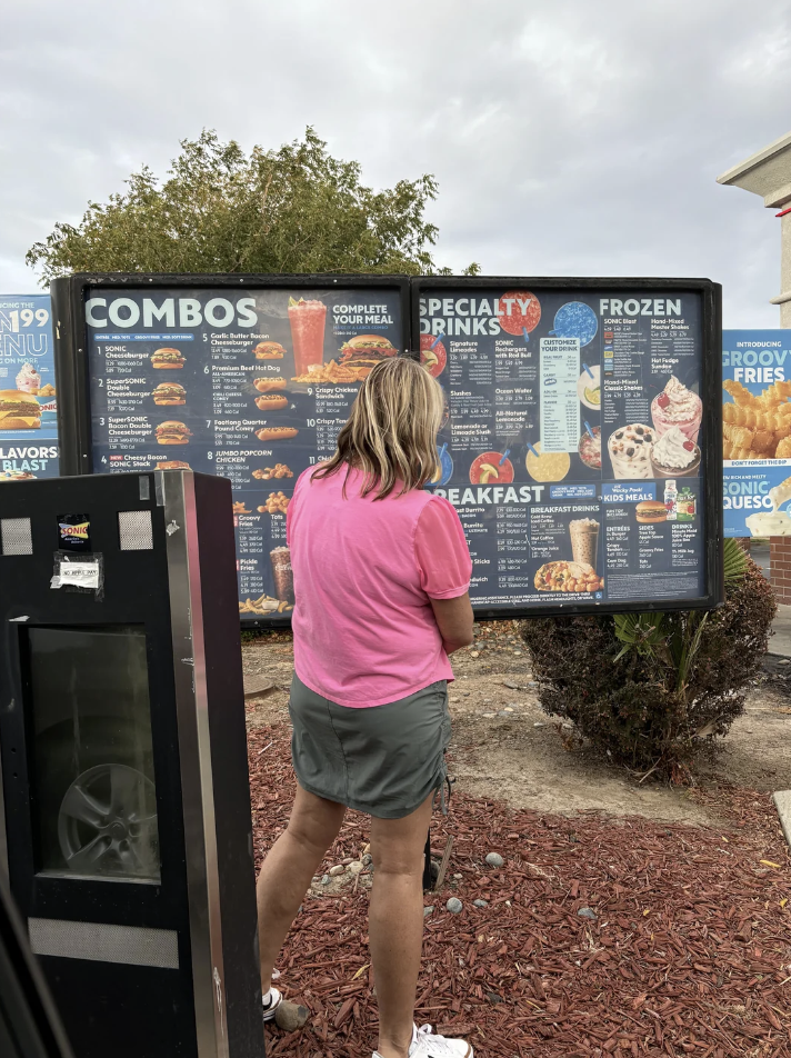 A person in a pink shirt and gray shorts stands at an outdoor fast food menu