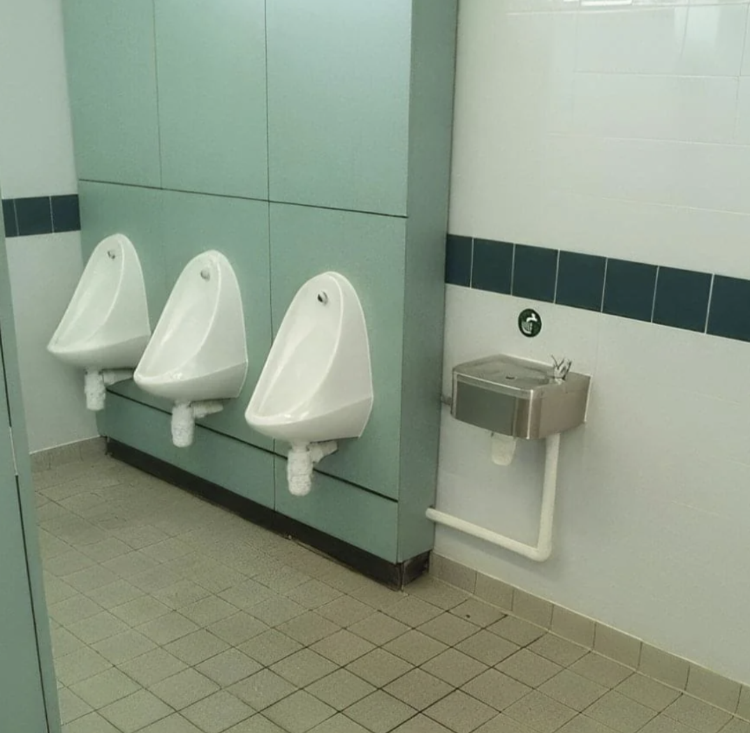 Three urinals are unusually placed close to a drinking fountain in a public restroom