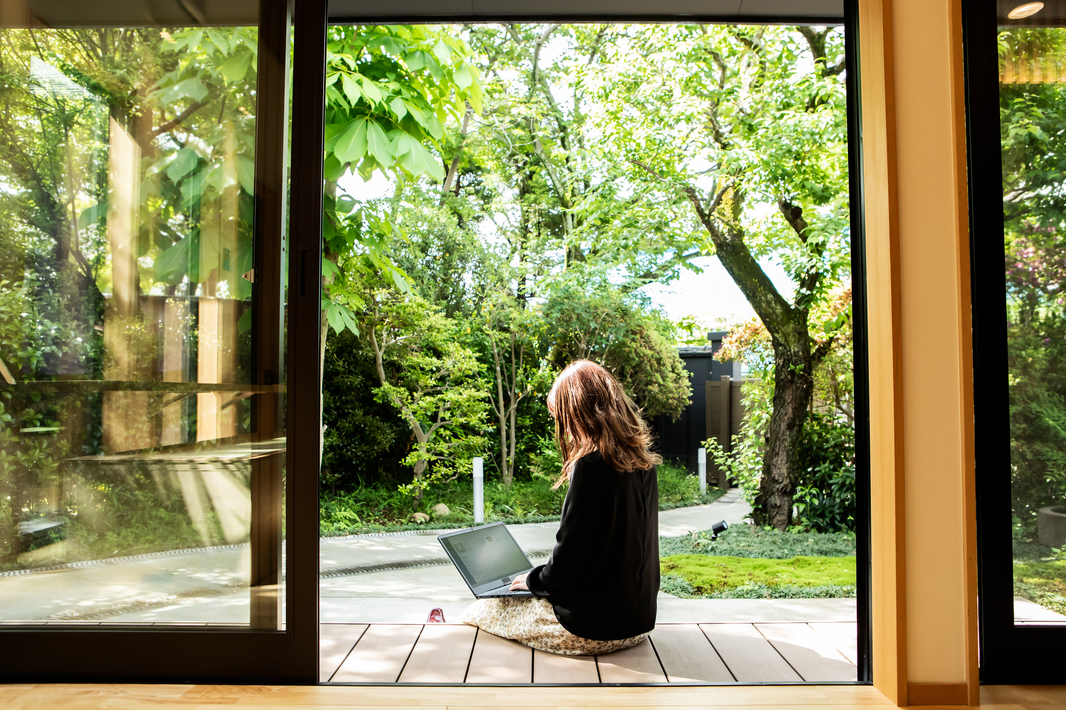 A person sits on a wooden deck with a laptop, looking out at a lush garden and trees through open glass doors
