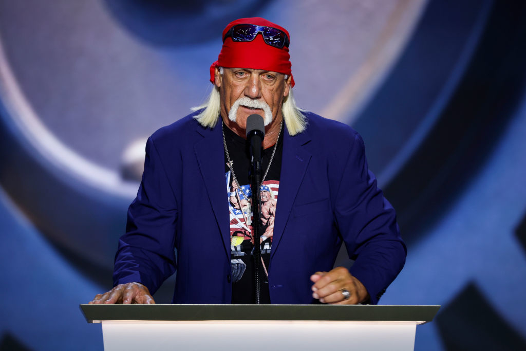 Hulk Hogan, wearing a bandana and blazer over a T-shirt, speaks at a podium