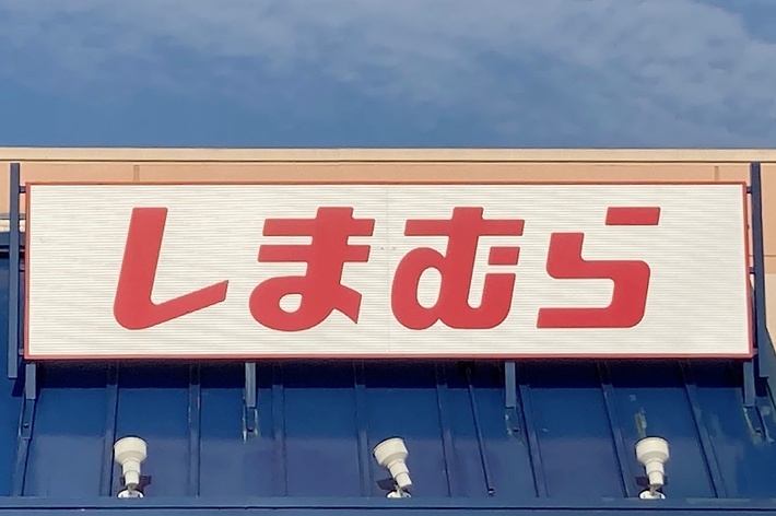 Storefront with a sign displaying Japanese text, &ldquo;しまむら,&rdquo; under a partly cloudy sky. Accessible parking sign visible at the bottom