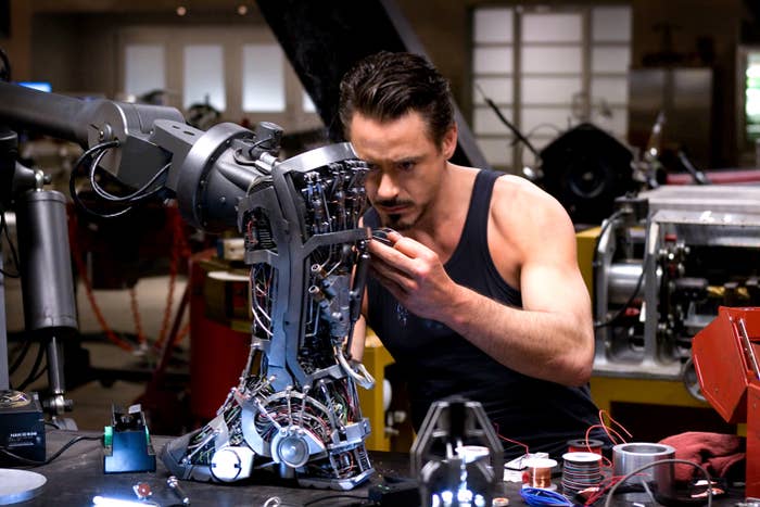 Robert Downey Jr. as Tony Stark, wearing a sleeveless shirt, works on a mechanical arm in a high-tech workshop from a scene in Iron Man