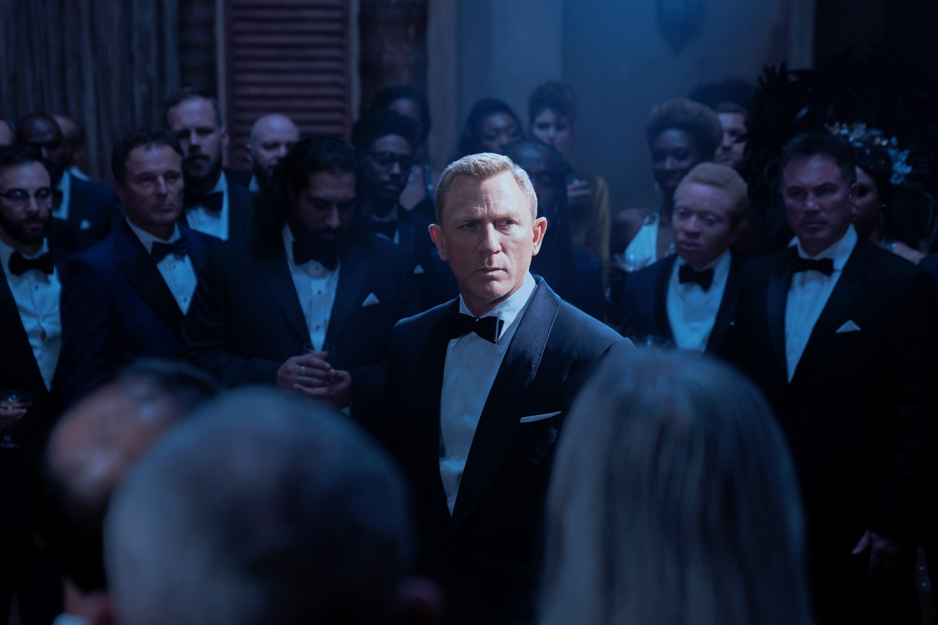 Daniel Craig, dressed in a classic tuxedo, stands amidst a crowd of formally dressed people at an elegant event in a scene from a movie