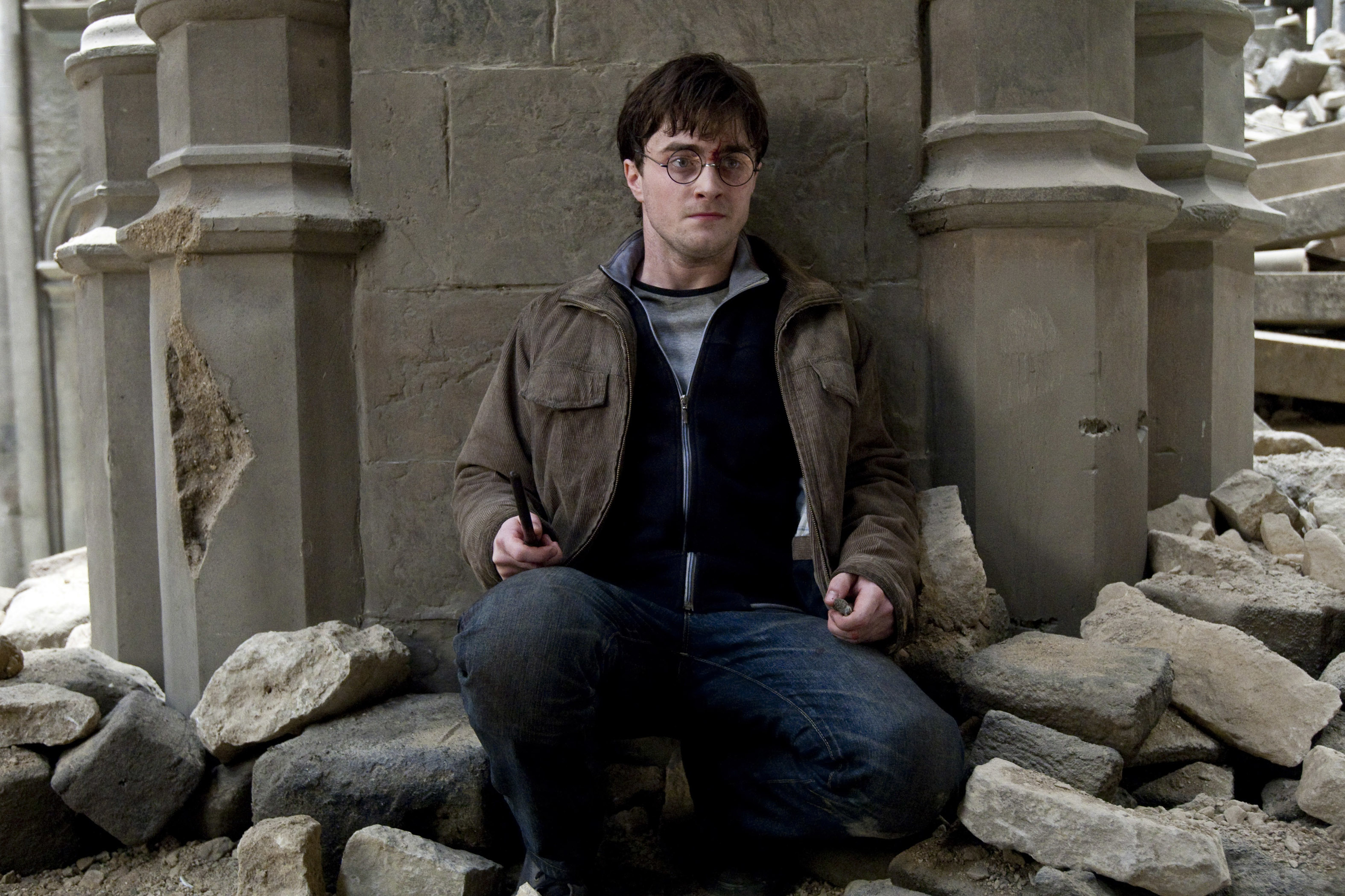 Daniel Radcliffe as Harry Potter sits surrounded by debris, holding his wand in both hands, wearing casual, rugged clothing
