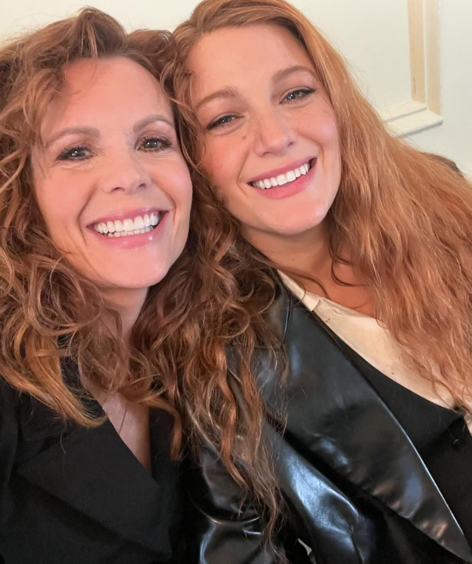 Two smiling women, both with curly hair, take a close-up selfie. They are dressed in casual chic attire