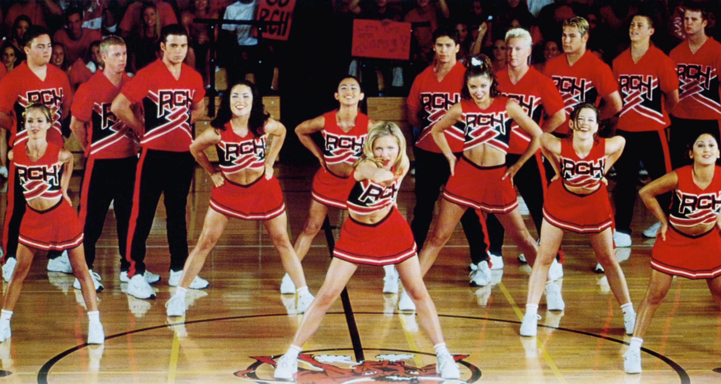 The cast of "Bring It On" dressed as cheerleaders in a scene performing a cheer routine in a gymnasium
