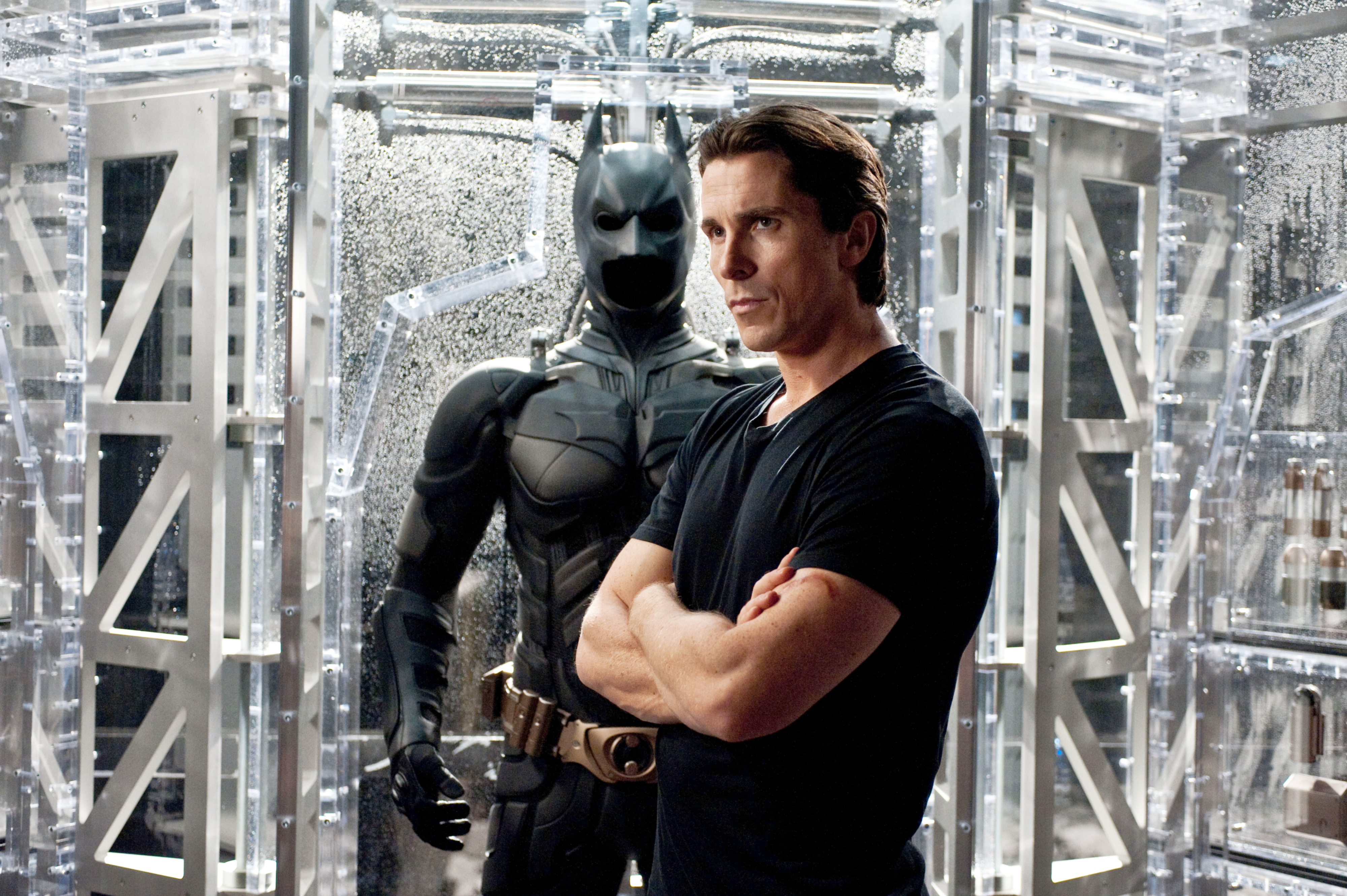 Christian Bale, in a black t-shirt, stands with arms crossed beside a Batman suit in a high-tech setting in a scene from a movie