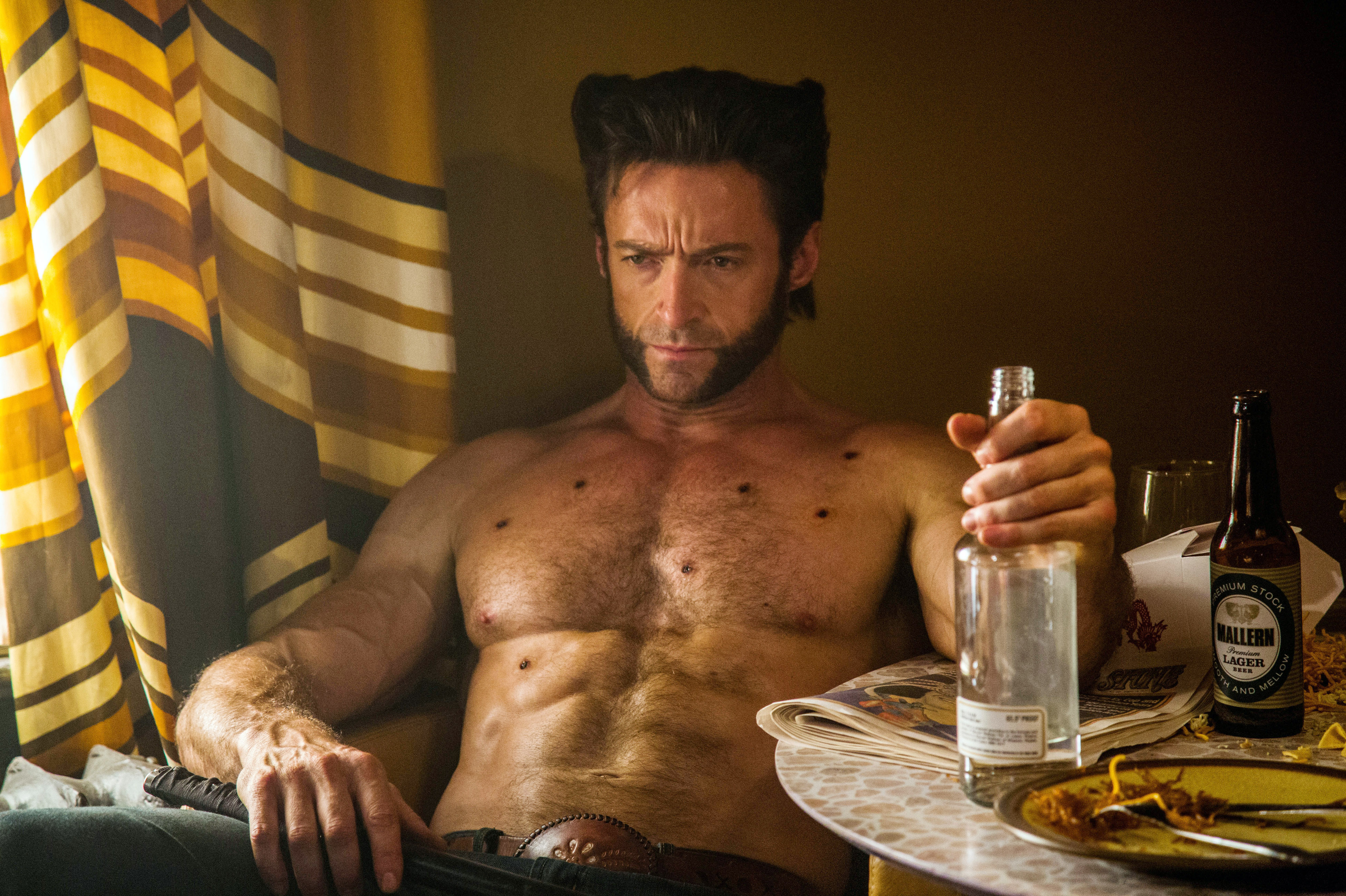 Hugh Jackman, shirtless and muscular, sits at a table with a bottle in hand. He appears contemplative. The table holds a beer bottle, glass, and some food
