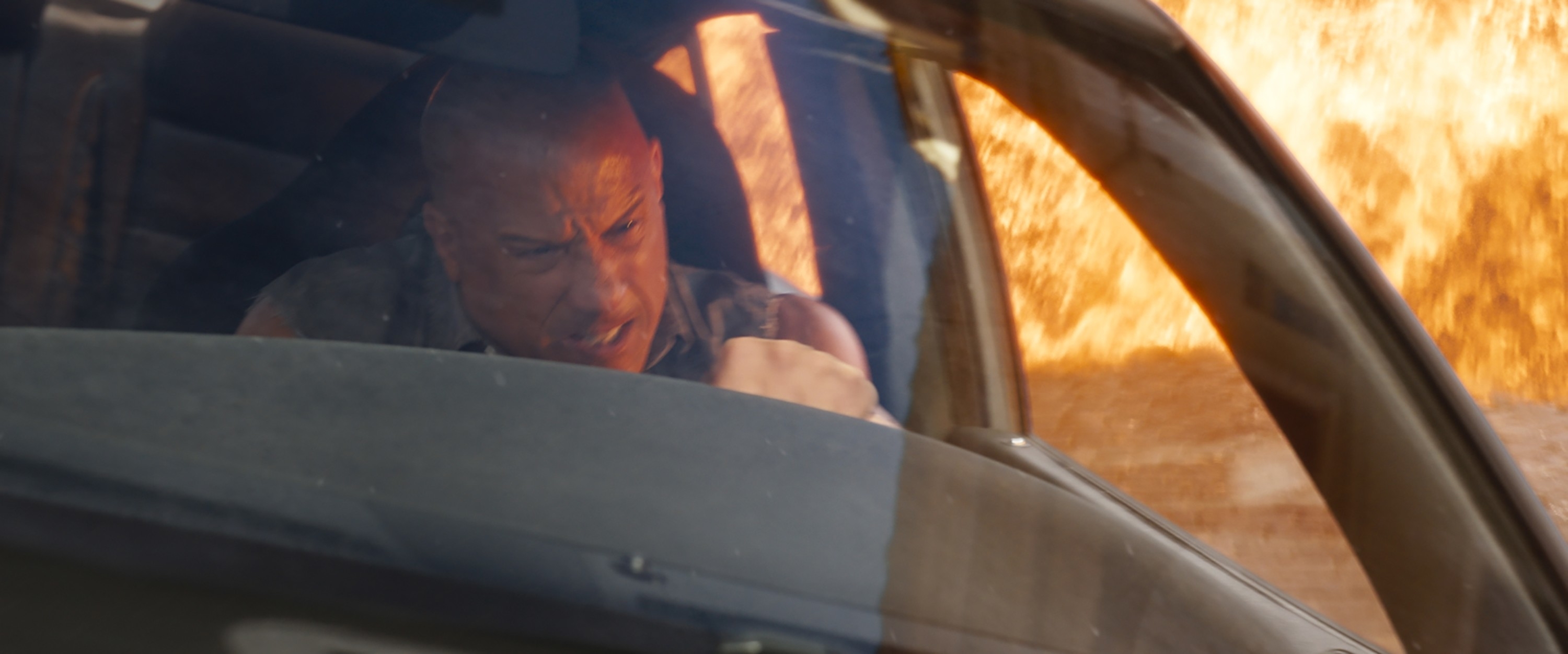 Vin Diesel intensely drives a car in an action scene with an explosion in the background