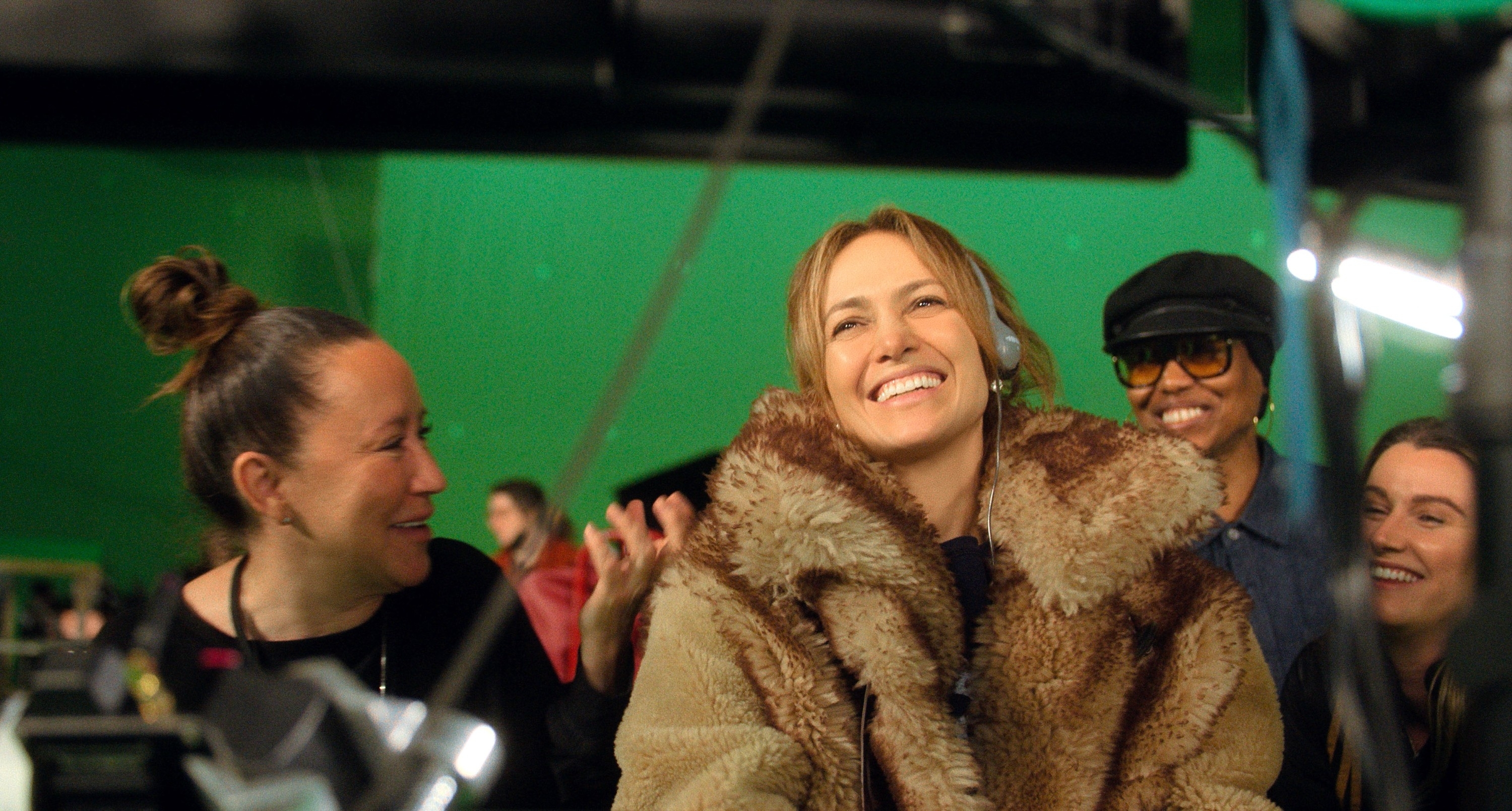 Jennifer Lopez smiling on a movie set, wearing a large fur coat, surrounded by crew members who are also smiling