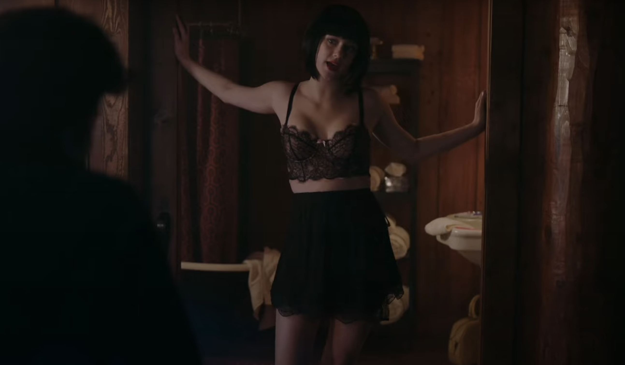 A woman in a lacy bra and skirt stands in a doorway facing a man whose back is to the camera in a dimly lit room