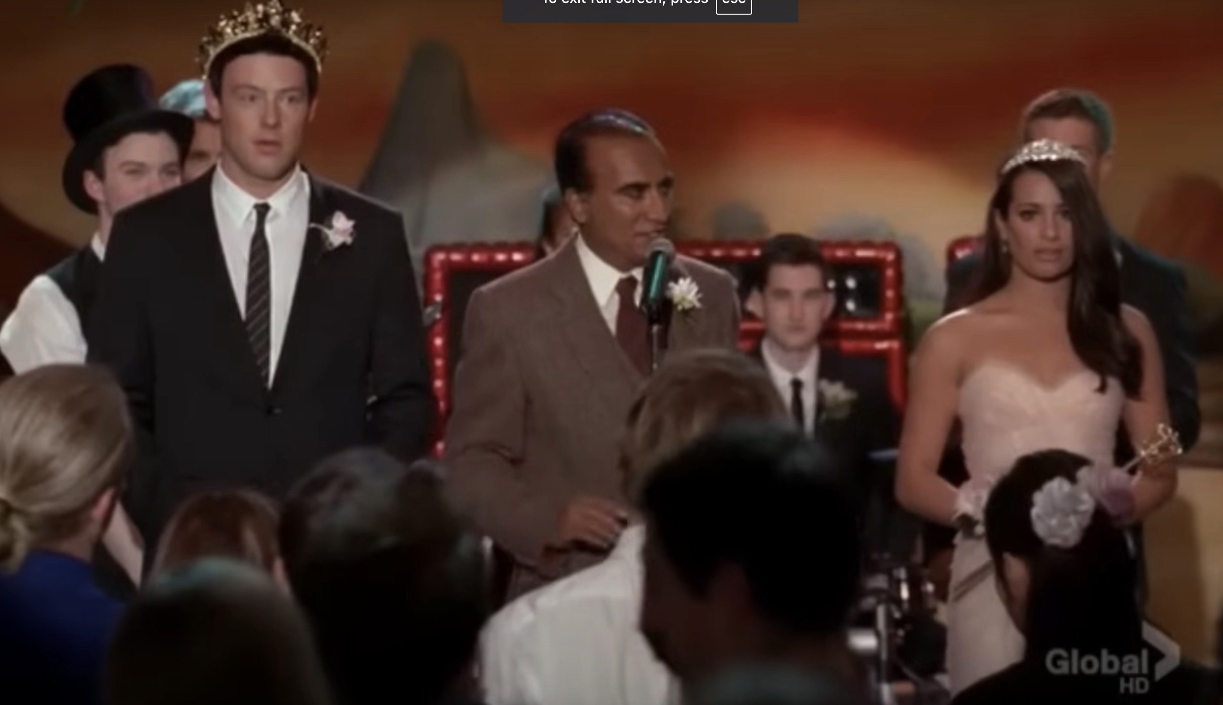 Cory Monteith, wearing a crown and suit, and Lea Michele, in a formal dress with a tiara, stand on stage at a formal event with a man speaking at a podium