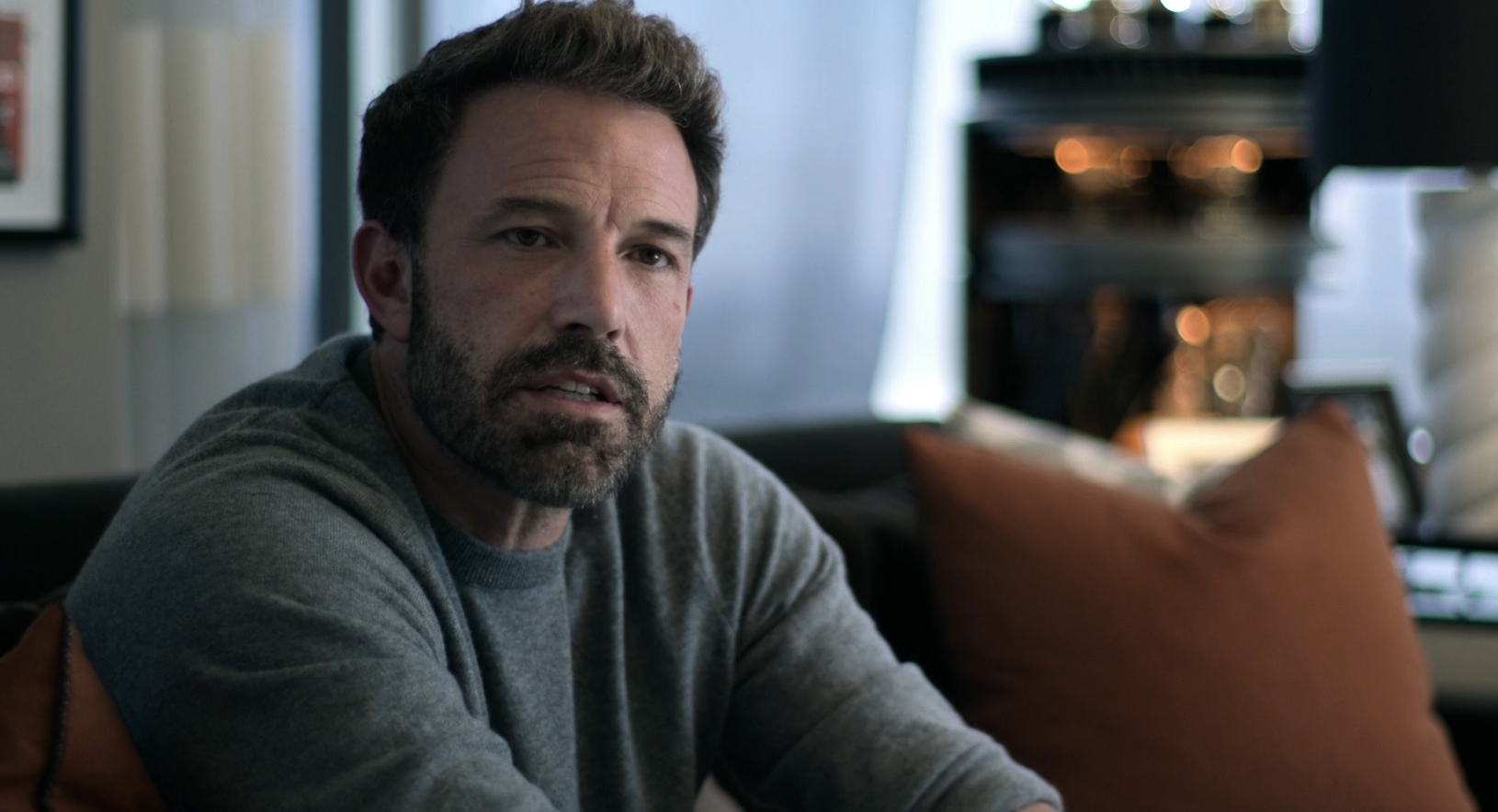 Ben Affleck sitting on a sofa, looking thoughtful, wearing a gray sweater