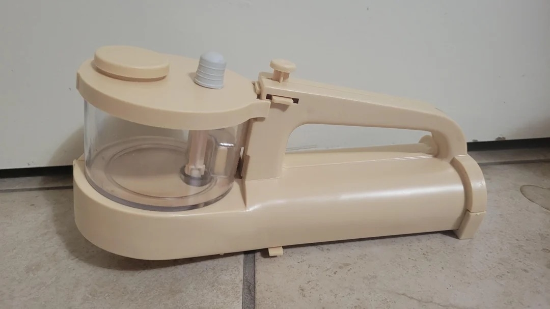 Handheld beige plastic item on a kitchen floor. The device has a clear bowl and handle for easy use
