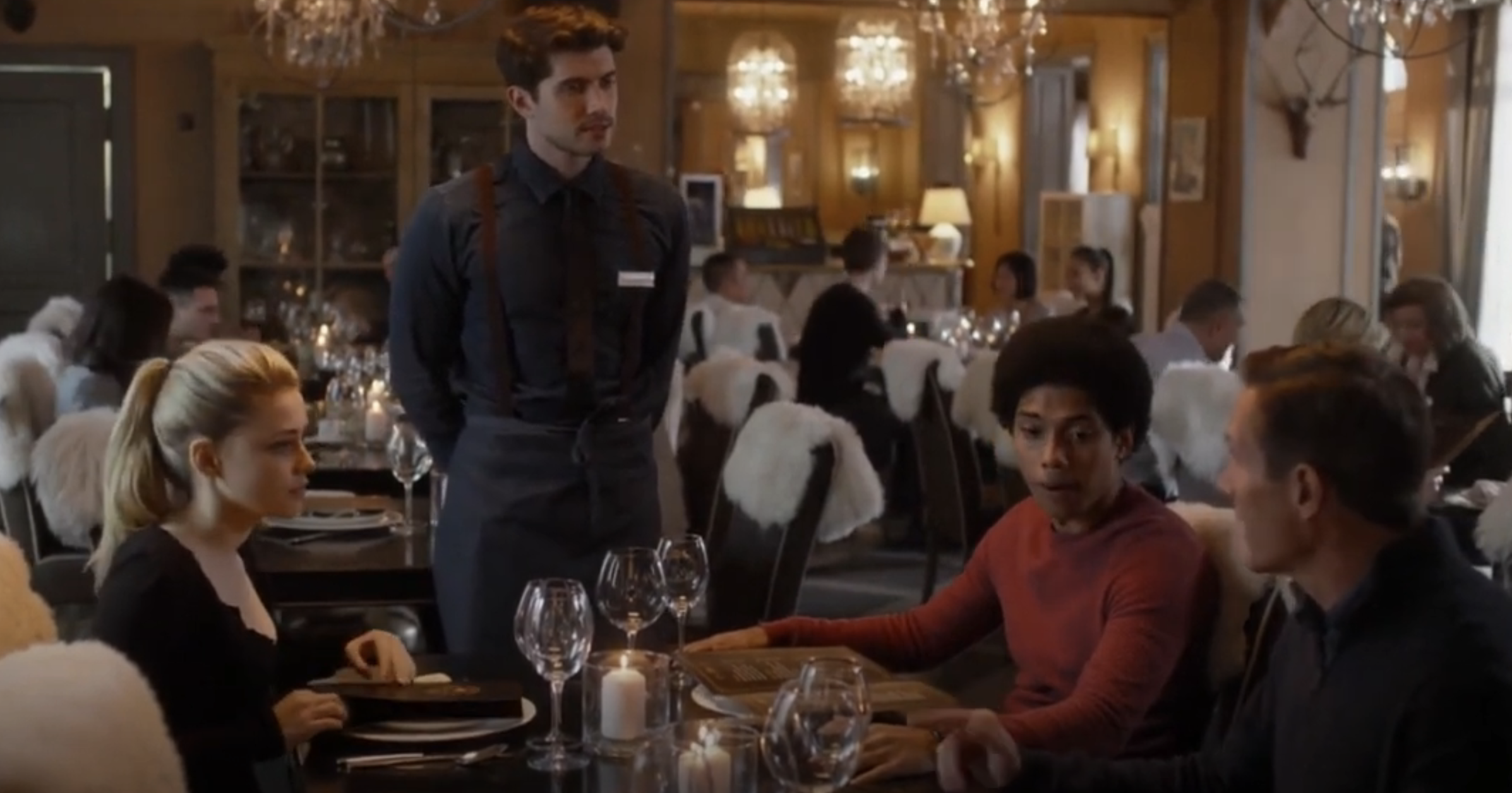 Three diners seated at a cozy restaurant table, being served by a waiter. The diners appear engaged in conversation