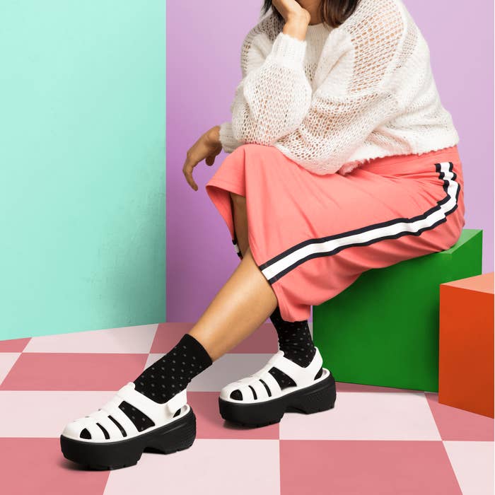 Woman seated in a modern, colorful room wearing a white sweater, pink track pants with black and white stripes, black polka dot socks, and white platform sandals