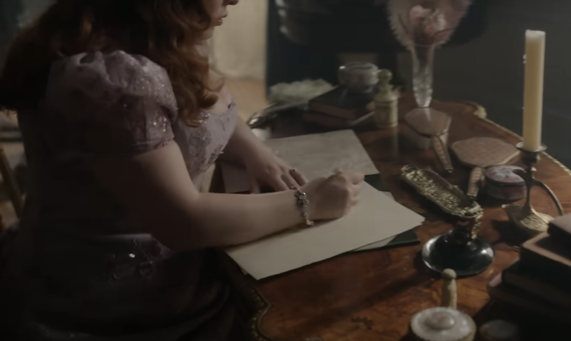 A woman in an ornate dress writes a letter with a quill on a wooden desk, surrounded by Victorian-era objects including candles, books, and a handheld mirror