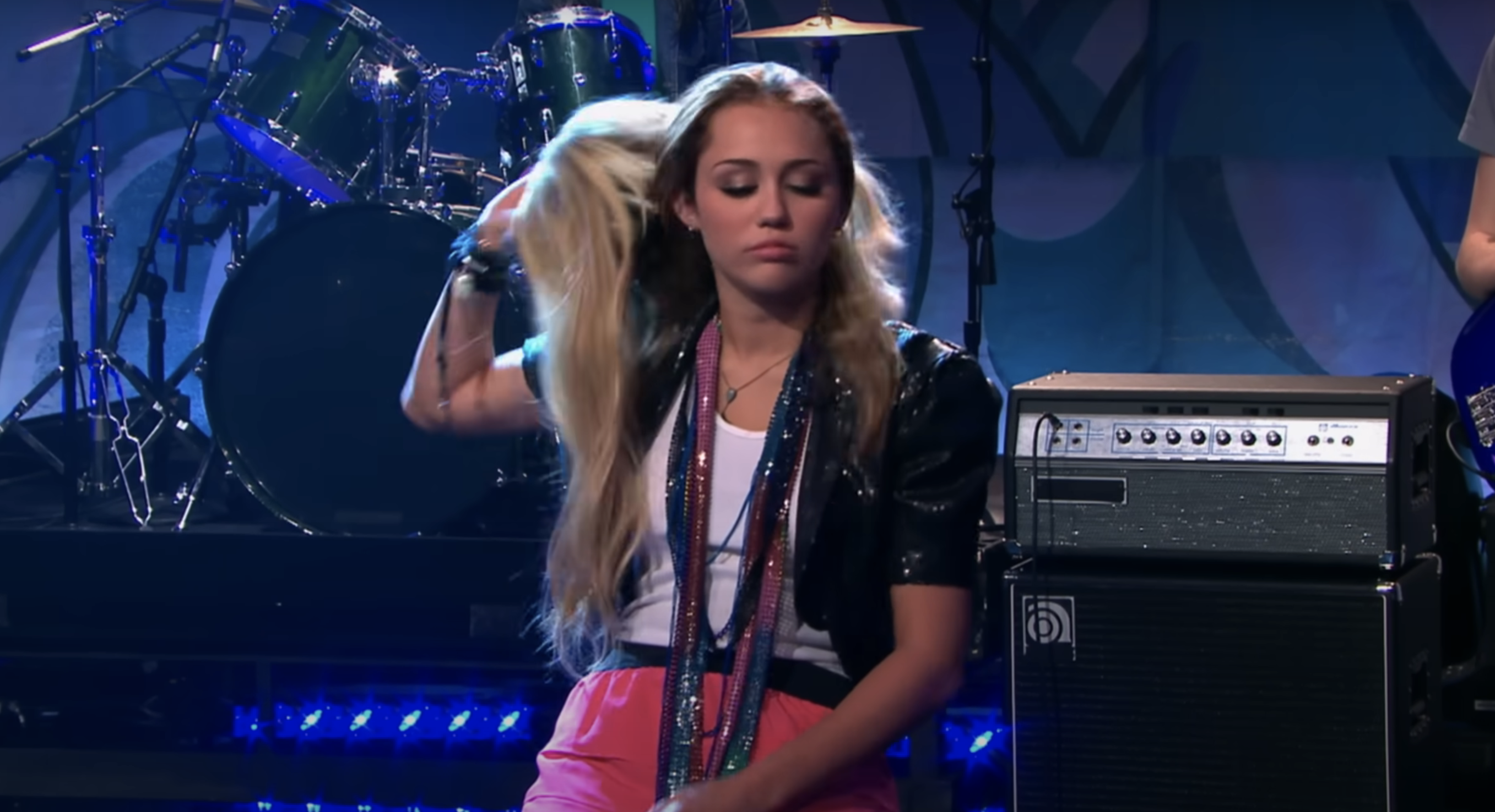 Miley Cyrus performing on stage, flipping her long hair with a serious expression. A drum set and an amplifier are visible in the background