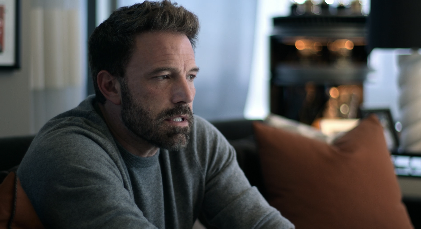 Ben Affleck wears a casual sweater, seated indoors, appearing thoughtful. He has a trimmed beard and mustache. The background includes pillows and home decor