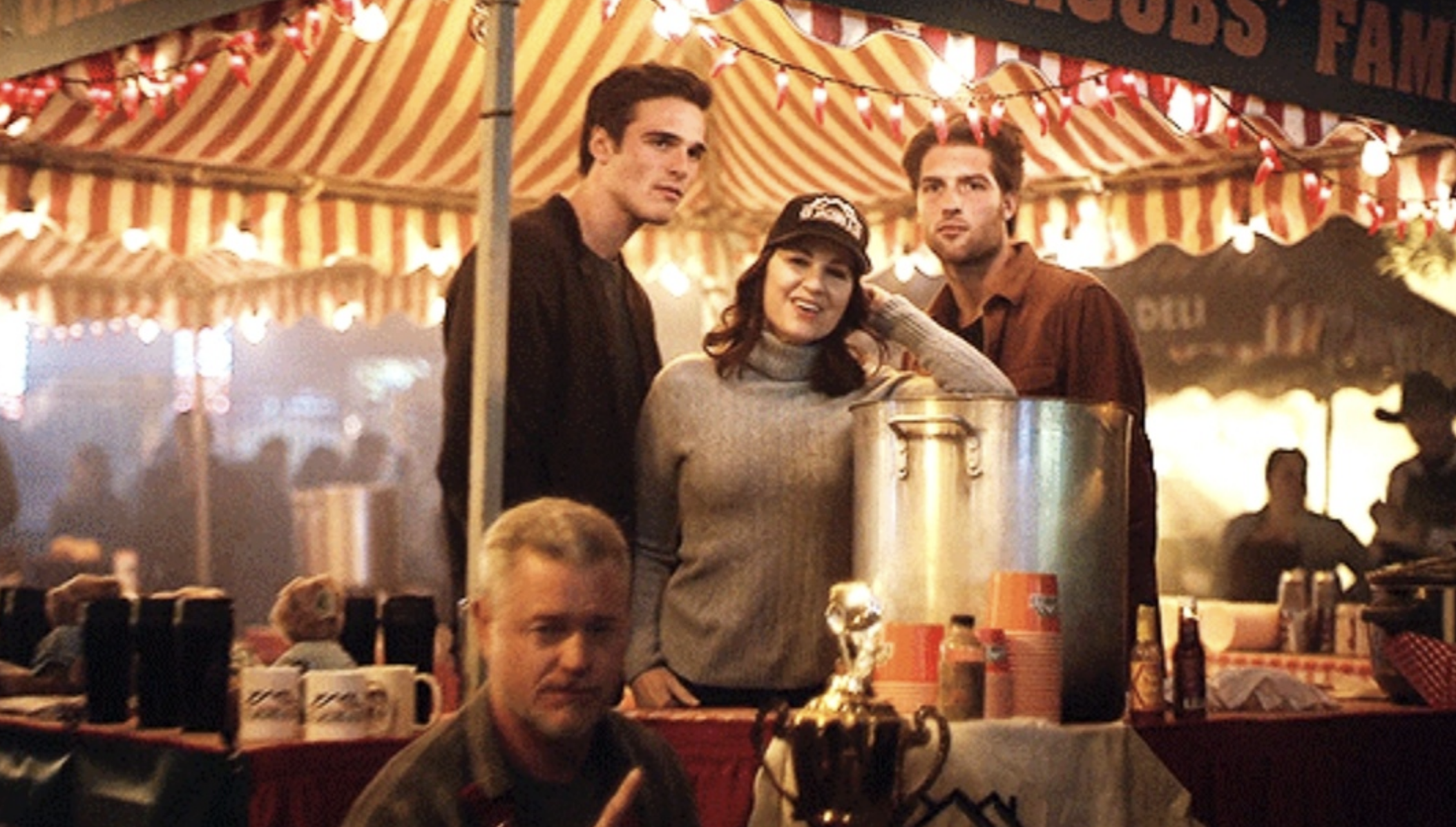 Four individuals in front of a carnival booth with a large cooking pot and trophy; three standing, one seated (names unknown). Patrons present behind them