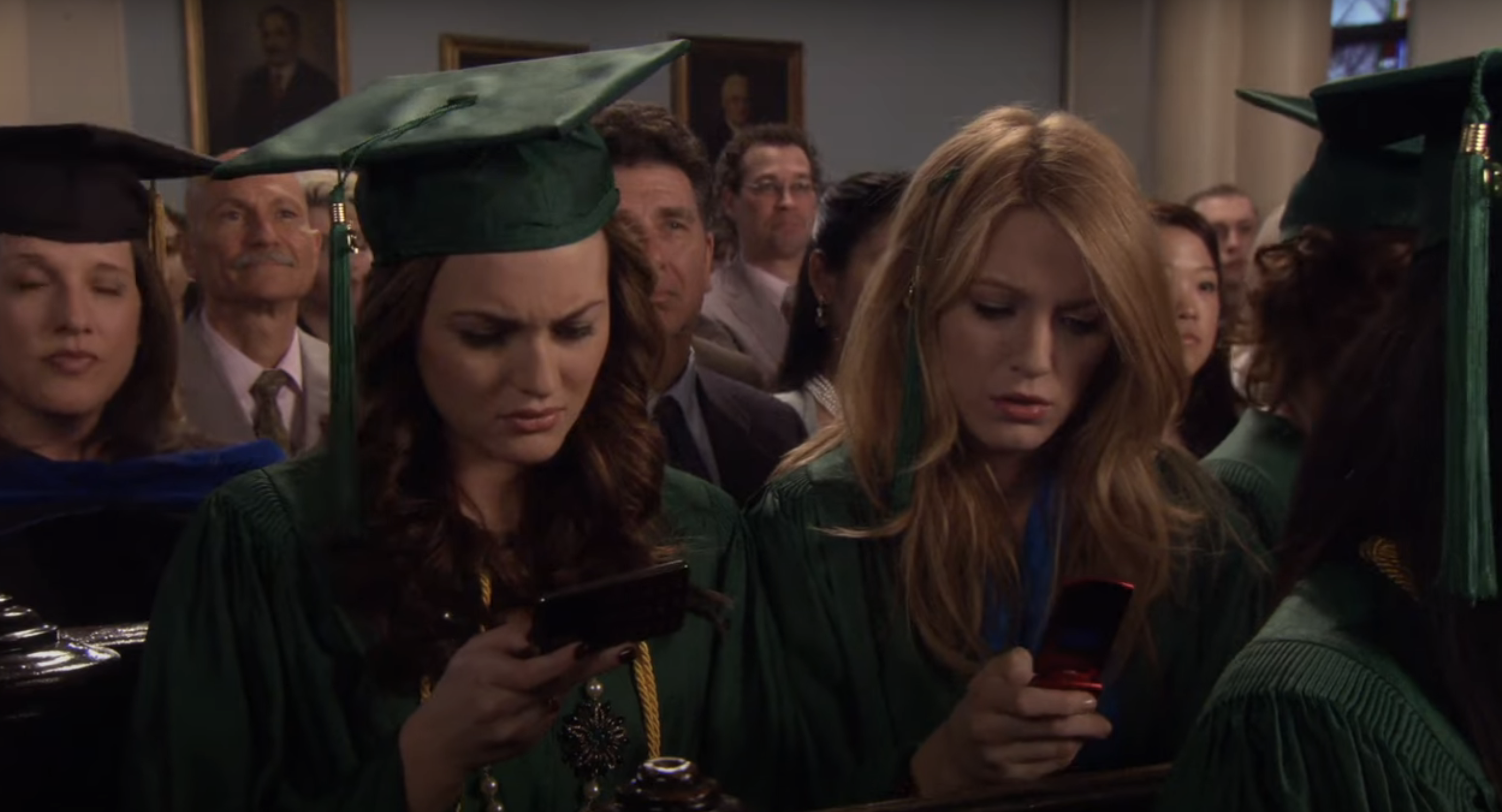 Leighton Meester and Blake Lively, dressed in green graduation gowns and caps, using their phones at a graduation ceremony